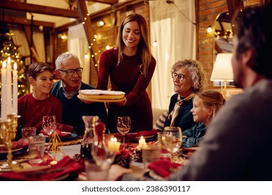 Happy woman serving Christmas pie to her extended family at dining table.