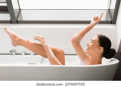 Happy Woman Shaving Legs Using Safety Razor In Bathtub, Enjoying Depilation Routine Taking Bath In Modern Bathroom At Home. Hair Removal And Bodycare Cosmetics Concept. Side View Shot - Powered by Shutterstock