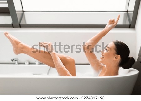 Happy Woman Shaving Legs Using Safety Razor In Bathtub, Enjoying Depilation Routine Taking Bath In Modern Bathroom At Home. Hair Removal And Bodycare Cosmetics Concept. Side View Shot