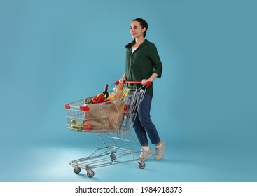 Happy woman with shopping cart full of groceries on light blue background