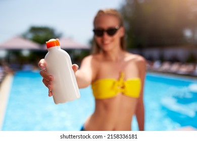 Happy woman shows suntan or sunscreen cream white bottle over the blurred swimming pool background. Tanned slim girl wearing yellow bikini swimwear and holds sun lotion. White bottle with copy space.