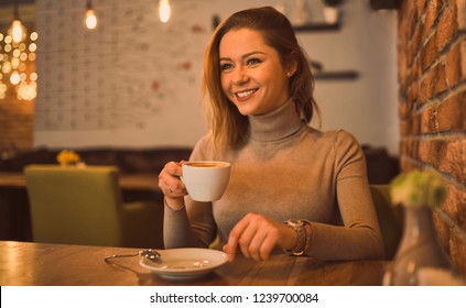 Happy woman sitting in a cafe, smiling, holding a cup of coffee. On the table is a plate with croissant. Looks ahead. - Shutterstock ID 1239700084