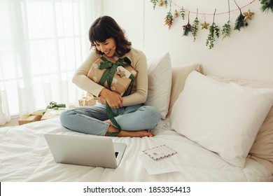 Happy Woman Sitting On Bed In A Decorated Room With Christmas Presents Doing Video Call. Woman Talking To Family And Friends On Video Call Using Laptop Computer.