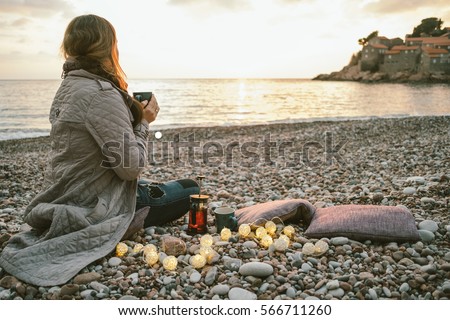 Happy woman sitting on cold beach with cup of tea with lights at sunset, Montenegro