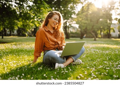 Happy woman sitting on a green clearing with a laptop. Portrait of a young freelance woman working outdoors. Online, technology concept. - Powered by Shutterstock