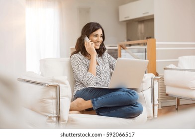 Happy Woman Sitting On Sofa With Laptop And Talking On Phone At Home. Young Successful Businesswoman Working From Home While Talking At Phone. College Student Studying On Laptop And Using Phone.