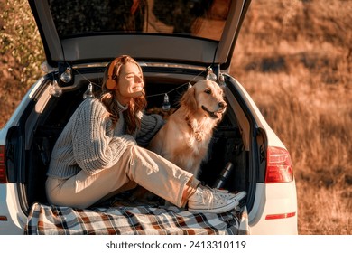 Happy woman sitting in the open trunk of a car with her dog on a warm blanket, listening to music on headphones. Woman traveling with pet.