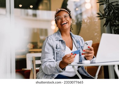 Happy woman sitting in a café and using her smartphone for online shopping, paying with a credit card. Young woman enjoying the convenience of mobile banking and digital transactions. - Powered by Shutterstock