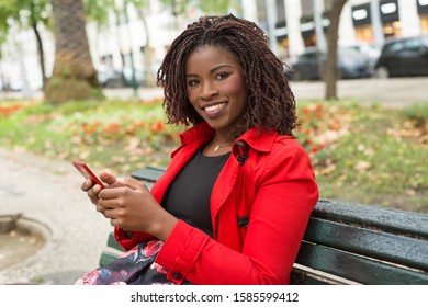 Happy woman with smartphone smiling at camera. Cheerful young African American woman sitting on bench and using cell phone. Technology concept - Shutterstock ID 1585599412