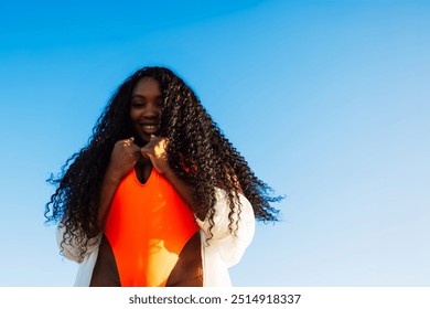 Happy woman smiles on a beach with wind in her curly hair. Enjoying the sunny day and tropical colors. She exudes confidence and joy in her orange swimsuit - Powered by Shutterstock