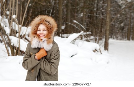 Happy woman smiles and wraps herself in warm clothes in winter forest on snow background. Wearing stylish fashion outfit khaki parka coat jacket with fur hood