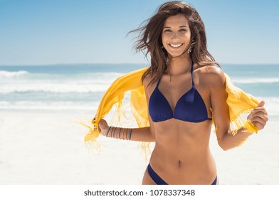 Happy woman smiling and having fun at beach. Summer portrait of young beautiful girl running on beach with a yellow scarf. Latin girl laughing and looking at camera with joy. 