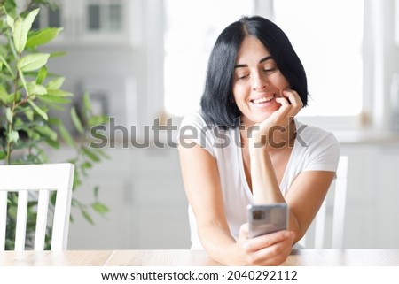 Happy woman smiling and holding smartphone in hands, young lady chatting in social networks online, watching funny videos in the internet, using mobile applications at home