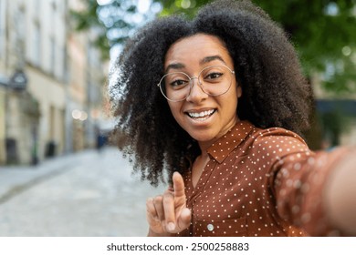 Happy woman smiling and making a video call outdoors using her phone. She is pointing at the camera, wearing glasses and a polka dot shirt, standing on a sunny street. - Powered by Shutterstock