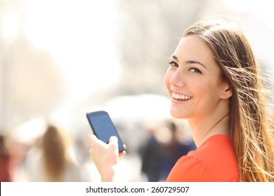 Happy woman smiling and walking in the street using a smartphone and looking at camera 