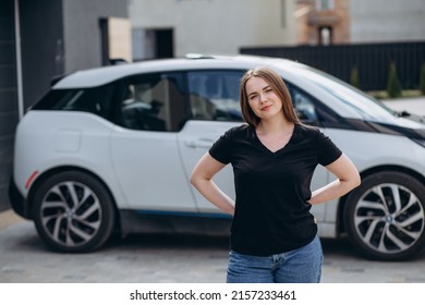 Happy Woman Standing Near Car, Leaning Elbow On Windshield And Putting Her Other Hand On Side Door Window. Girl Caressing Face Under Sunlight, Closing Eyes Against Charging Station And Solar Panel.