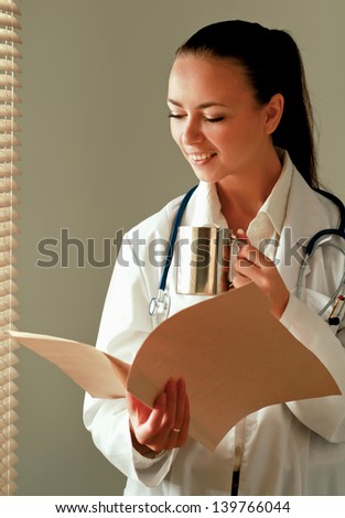 Happy woman is standing in the office near window with folder and holding cup
