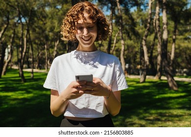 Happy woman standing on a city park while messaging with friend on smart phone. Redhead woman using phone or watching video or scrolling social media with big smiles.