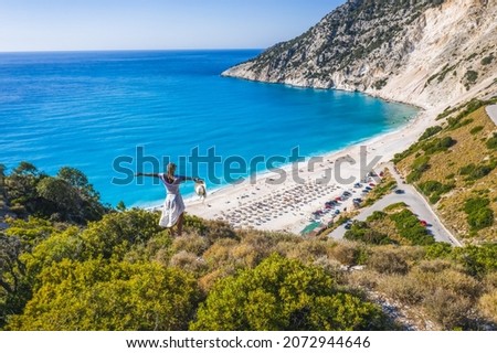 Happy woman standing on top of a rock, raising hands with an exciting feeling of freedom, looking at Myrtos Beach. Cephalonia island, Greece