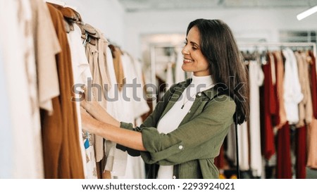 Happy woman stands in a fashion store, carefully choosing clothing items to buy. She browses through racks of stylish clothes, examining each one closely before making a decision.