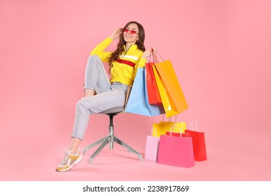 Happy woman in stylish sunglasses holding many colorful shopping bags on armchair against pink background - Powered by Shutterstock