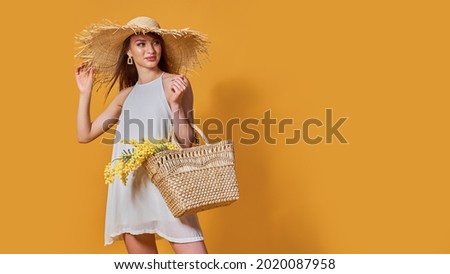Happy woman in summer white dress, straw hat with bunch of flowers in bag posing on yellow studio background.