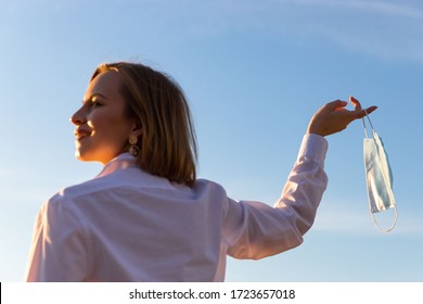 Happy woman takes off medical protective mask holds it on her finger on blue sky background, enjoys life, clean fresh air after Covid-19 pandemic, self-isolation. Quarantine is over. Soft focus