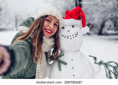 Happy woman takes selfie by snowman in Santa hat outdoors in snowy winter park. Christmas festive season. Fun activities - Powered by Shutterstock