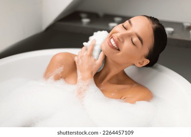 Happy Woman Taking Bath Rubbing Neck With Sponge And Foam Posing With Eyes Closed Enjoying Bathing In Modern Bathroom At Home. Bodycare And Wellness Concept. Selective Focus - Powered by Shutterstock