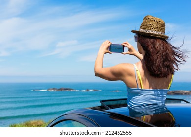 Happy woman taking photos to the sea with smartphone camera on car summer travel vacation to the coast. Brunette girl  leaning out vehicle sunroof towards the ocean. - Powered by Shutterstock