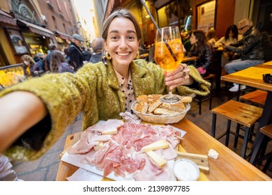 Happy Woman Taking Selfie Photo On Phone While Sitting With Meat Plate And Cocktail At Outdoor Bar In Bologna City. Concept Of Italian Gastronomy And Travel