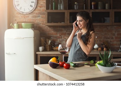 Happy Woman Talking On Phone And Drinking Wine While Cooking Healthy Food In The Loft Kitchen At Home On Sunny Day. Preparing Vegetable Salad.