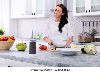 Happy Woman Talking To Voice Assistant While Slicing Cucumber With Kitchen Knife