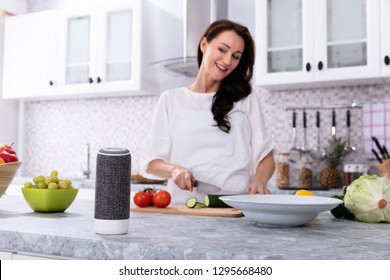 Happy Woman Talking To Voice Assistant While Slicing Cucumber With Kitchen Knife