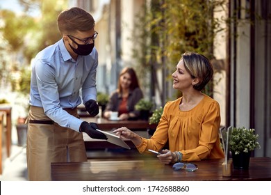 Happy woman talking to a waiter who is wearing protective face mask while choosing something from a menu on touchpad in a cafe.  - Powered by Shutterstock