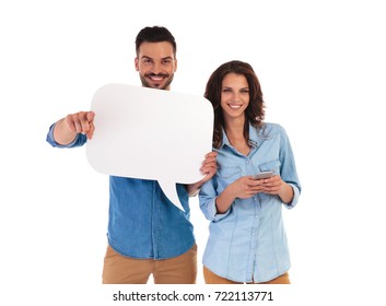 Happy Woman Is Texting On Phone While Man Is Holding A Speech Bubble And Points To The Camera On White Background