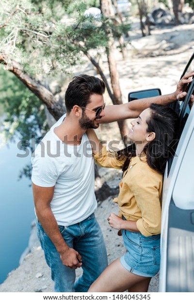 happy woman touching face of bearded man while standing\
near car 