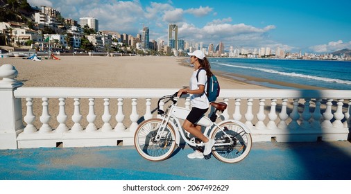 Happy Woman Tourist With Electric Bicycle Over Background Levante Beach Overlooking Panorama Of Benidorm City. Traveler Concept