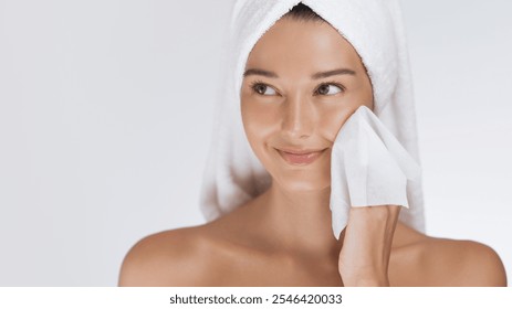 A happy woman with a towel wrapped around her head cleanses her face with a wipe, enjoying her skincare routine. Perfect for illustrating concepts of beauty, self-care, hygiene, and fresh skin care. - Powered by Shutterstock