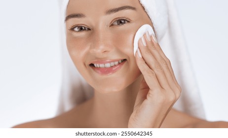 A happy woman with a towel wrapped around her head uses a cotton pad on her face, enjoying her skincare routine. Ideal for illustrating beauty, self-care, hygiene, and facial care concepts. - Powered by Shutterstock