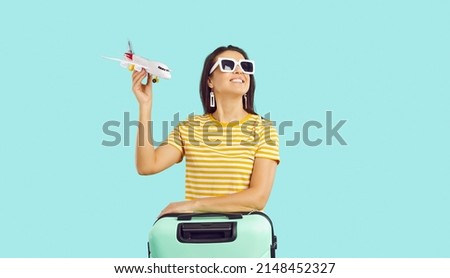 Happy woman traveler in sunglasses with suitcase hold airplane model in hands isolated on green studio background. Smiling girl with baggage ready for summer vacation. Travel and tourism.
