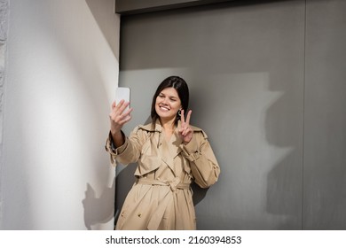 Happy Woman In Trench Coat Taking Selfie And Showing Peace Sign Near Wall