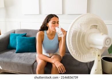 Happy woman trying to cool down drinking an ice cold drink and turning on the electric fan at home - Shutterstock ID 2146836739
