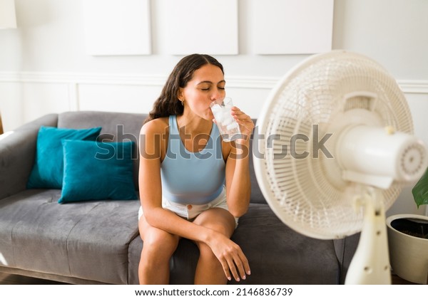 Happy woman trying to cool down\
drinking an ice cold drink and turning on the electric fan at\
home