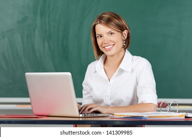 Happy woman typing on a laptop sitting at a table in front of green chalkboard smiling at the camera