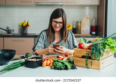 Happy Woman Using Mobile Phone While Making Healthy Lunch In The Kitchen