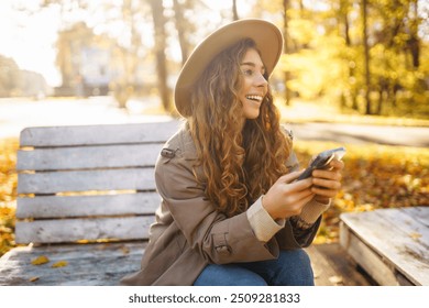 Happy woman using mobile phone on a bench in a city autumn park, young woman smiling while use application. Concept of vacation, blogging, technology, weekend. - Powered by Shutterstock