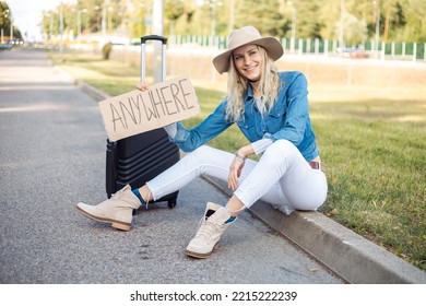 Happy Woman Wait Passing Car Sitting With Suitcase And Cardboard Poster On Roadside Of Empty Road. Blonde Lady In Hat Escape From City By Auto Stop To Go Anywhere. Travelling, Hitchhiking, Vacations.