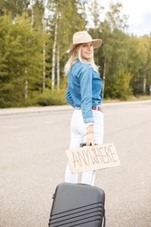 Happy Woman Walk Along Empty Road With Cardboard Poster And Suitcase, Back View. Lady In Hat And Denim Outfit Escape From City By Auto Stop To Go Anywhere. Travelling, Freedom, Hitchhiking, Vacation.
