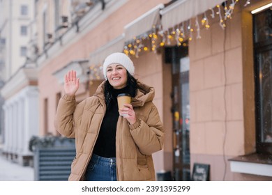 A happy woman walks through the city in winter with a cup of coffee and waves her hand - Powered by Shutterstock